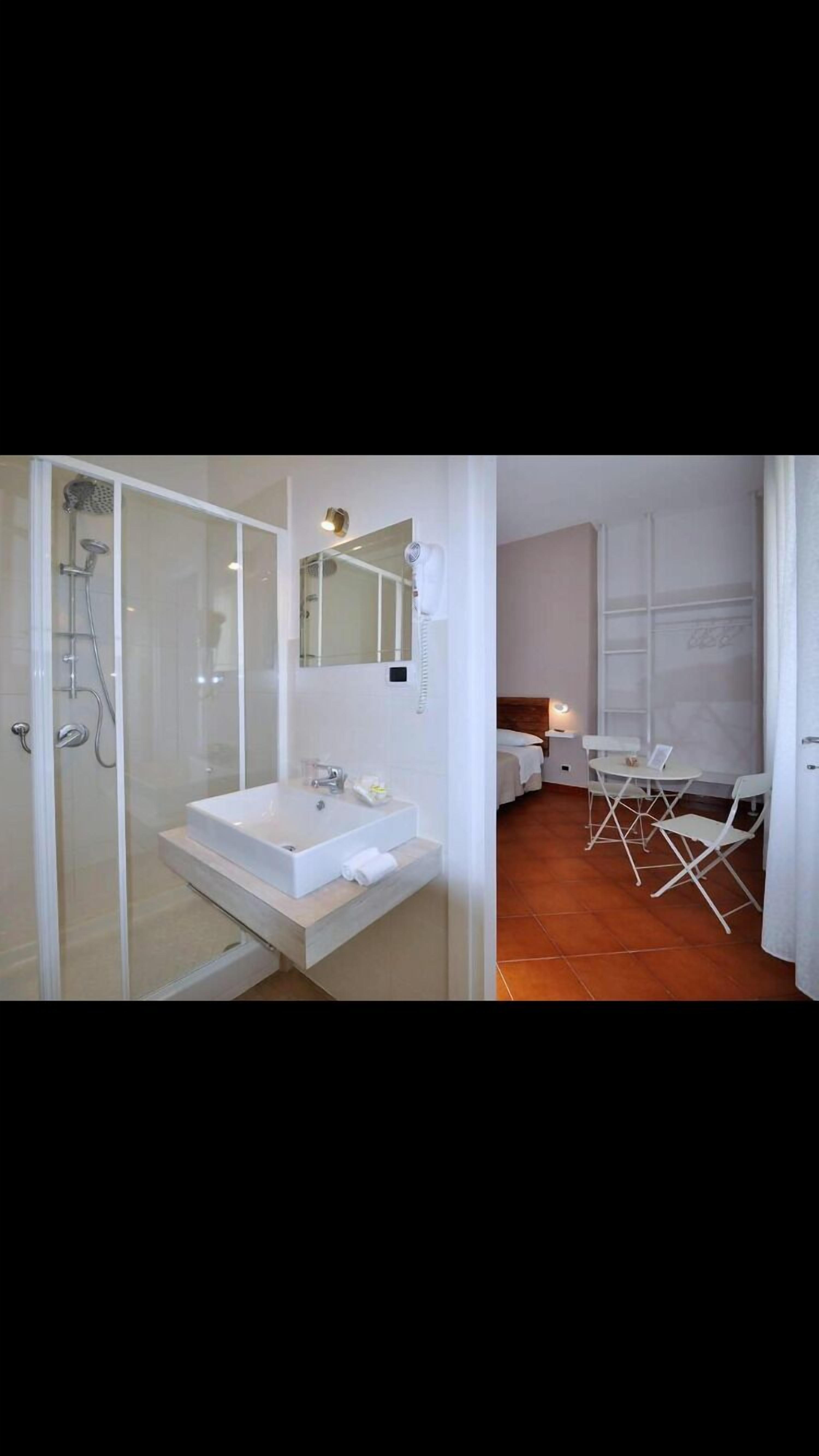 Un baño con ducha de vidrio, un lavamanos y un espejo. Junto a él, un dormitorio con cama, mesita de noche y una pequeña mesa con sillas.