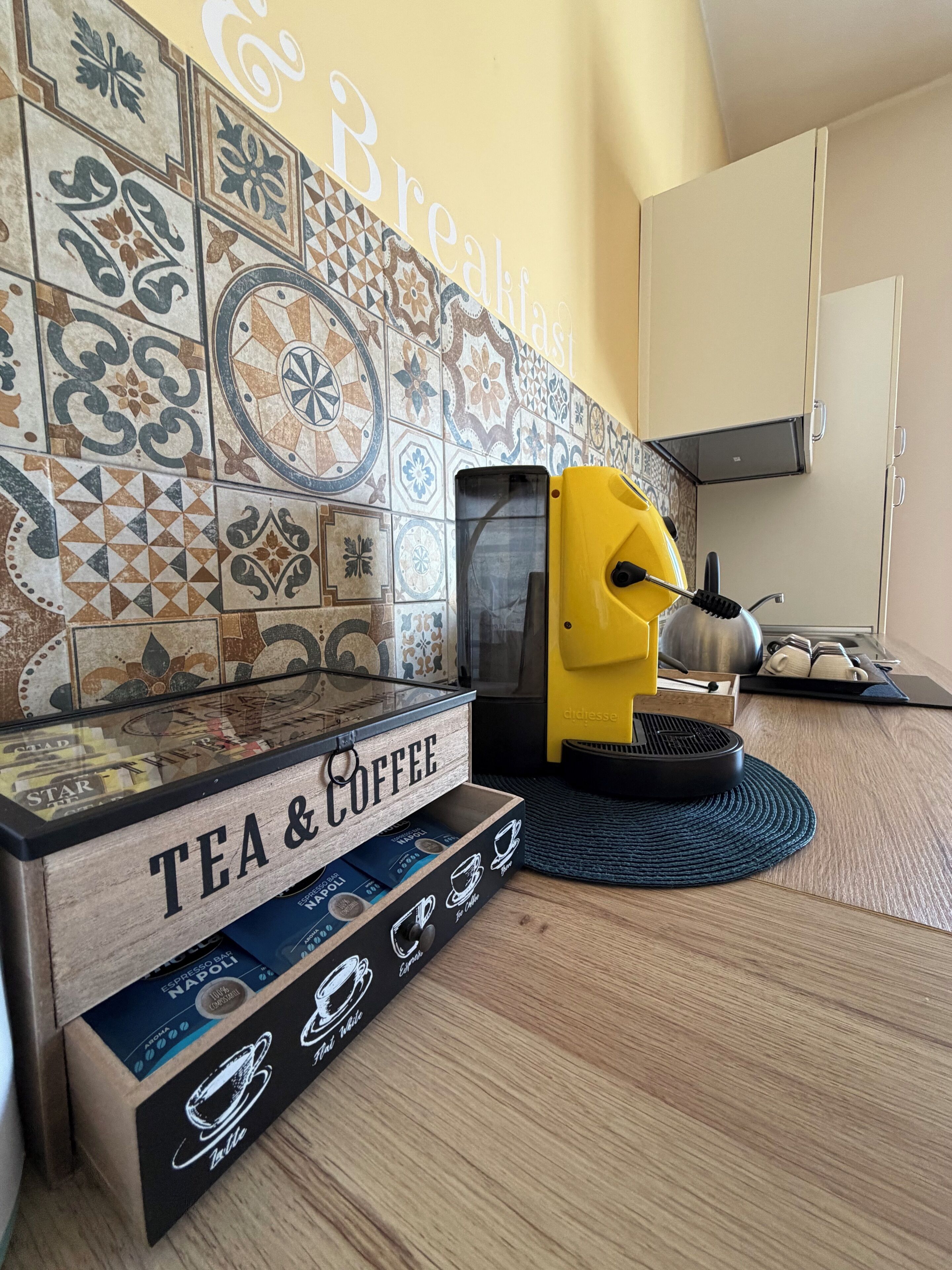 Une cuisine équipée d’une machine à café, d’une boîte contenant du thé et du café, et d’un mur sur lequel est affiché un panneau indiquant « Petit-déjeuner ».