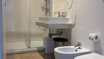 Un baño moderno con cabina de ducha de vidrio, lavabo con espejo, dos inodoros y dispensador de papel higiénico.