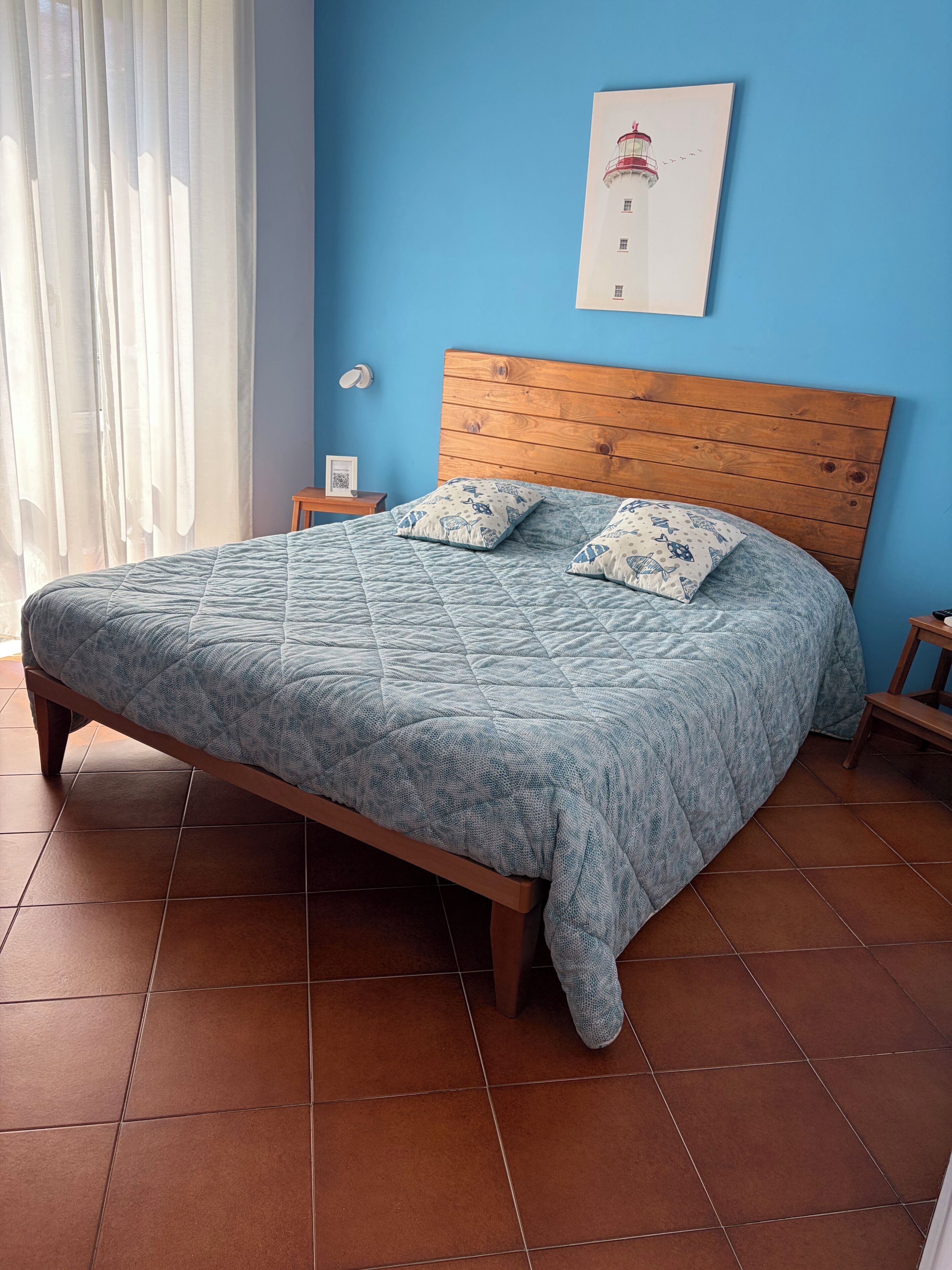 A bedroom with a wooden headboard, a quilted bedspread, two pillows, a nightstand, a framed picture on the wall, and a window with curtains.