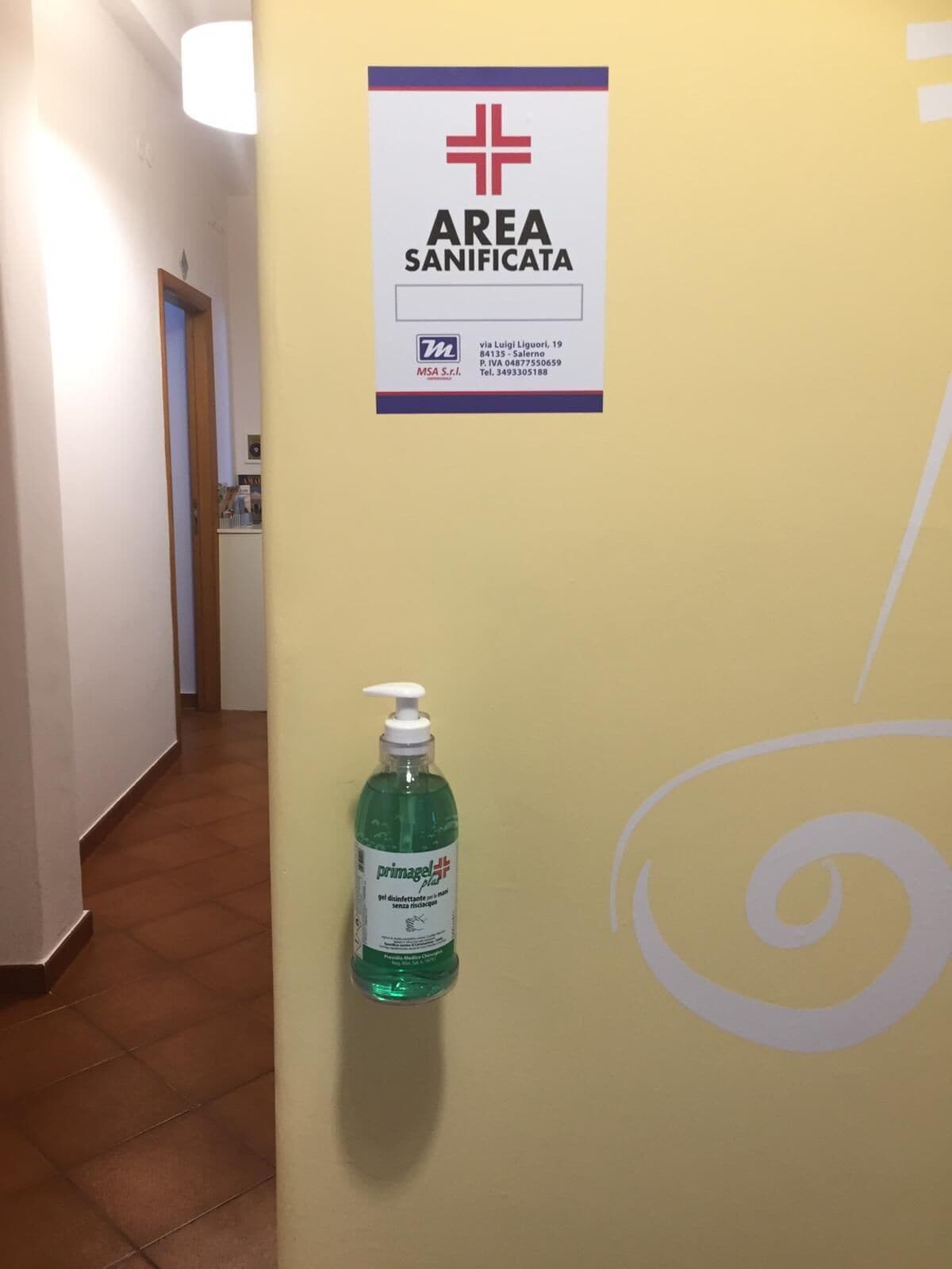 A hallway with a sign indicating an 'AREA SANIFICATA' and a hand sanitizer dispenser.
