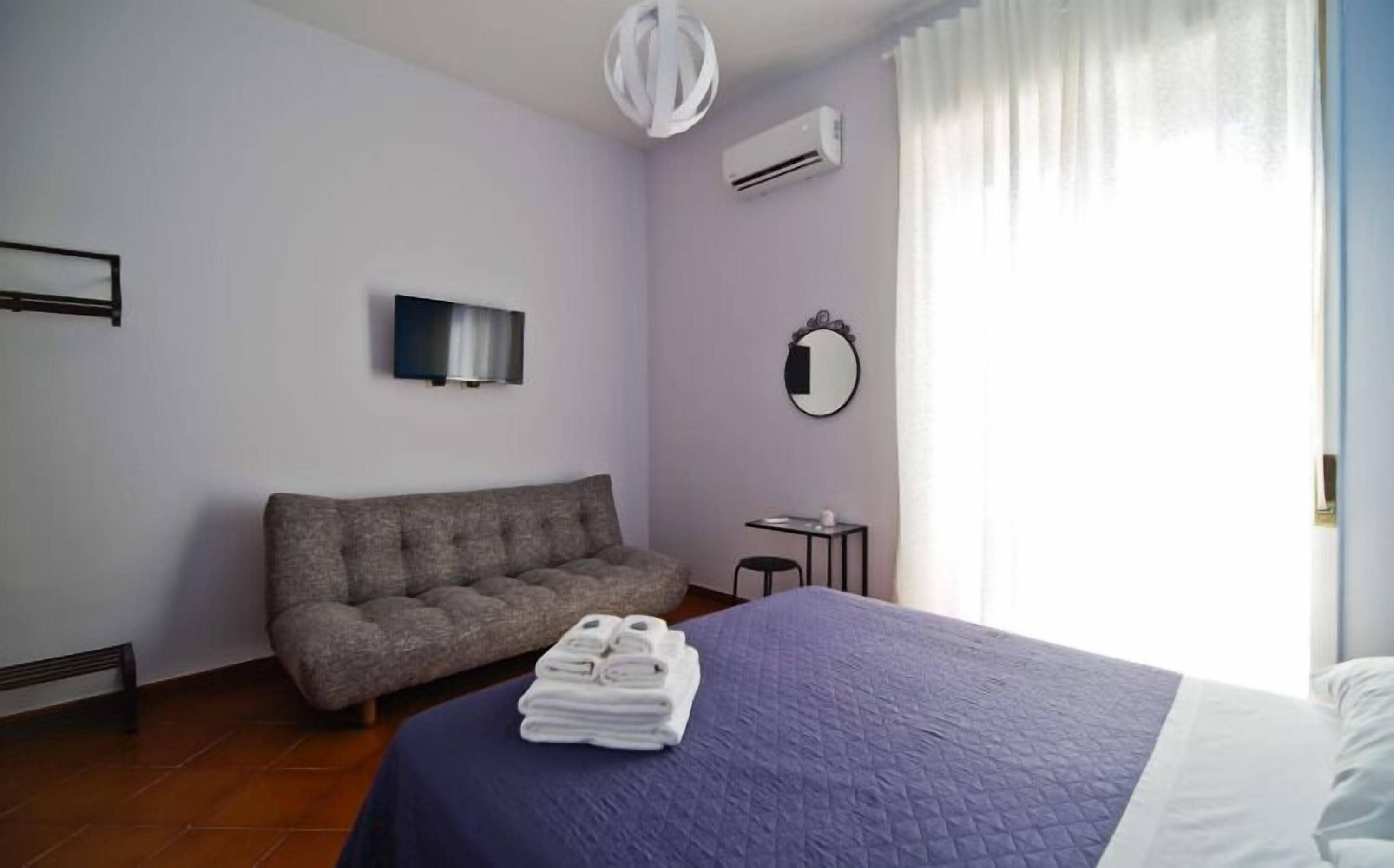 A bedroom with a bed, a sofa, a TV, an air conditioner, and a mirror.