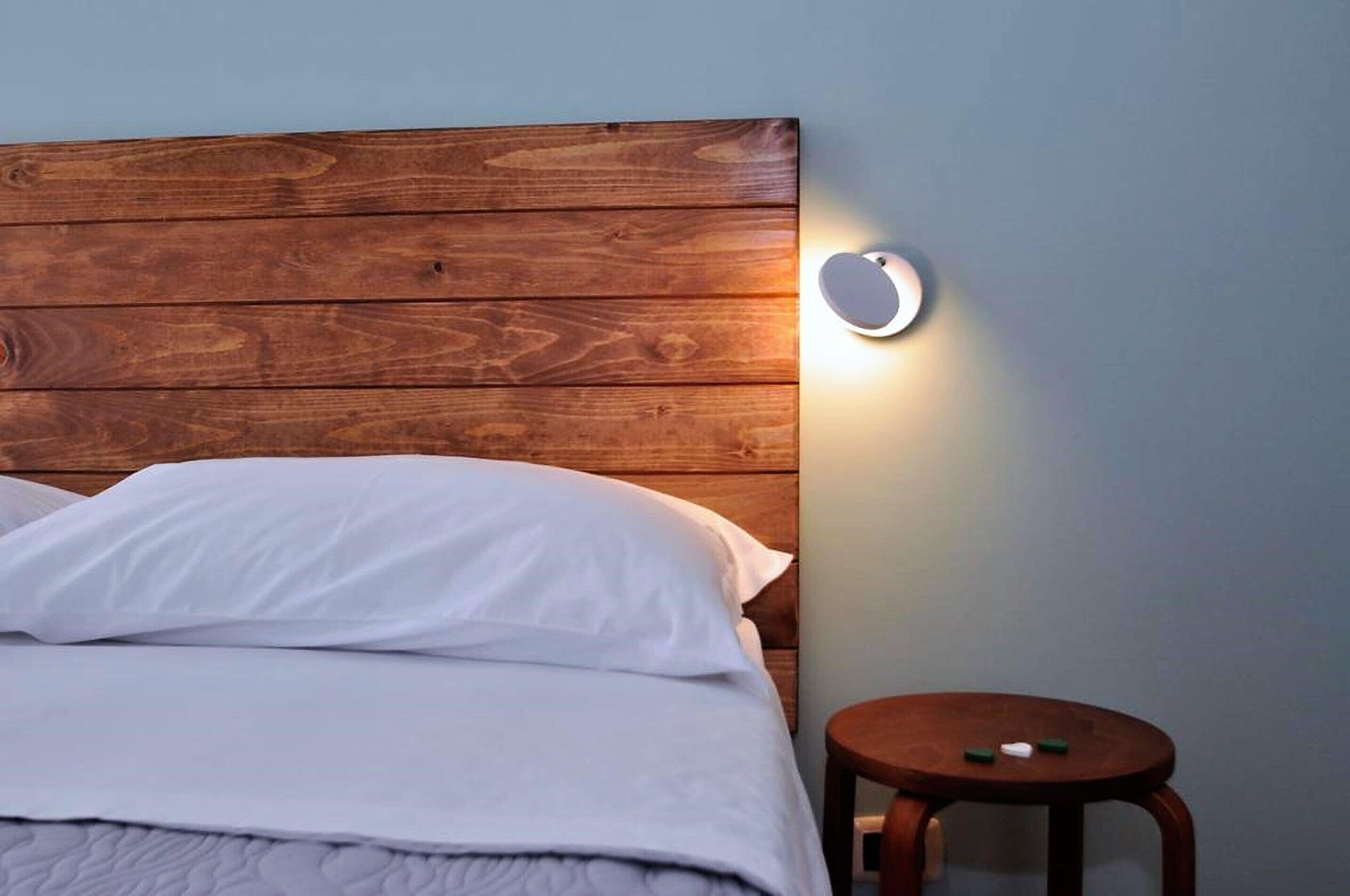 A bed with white bedding, a wooden headboard, and a round nightstand with a small lamp.
