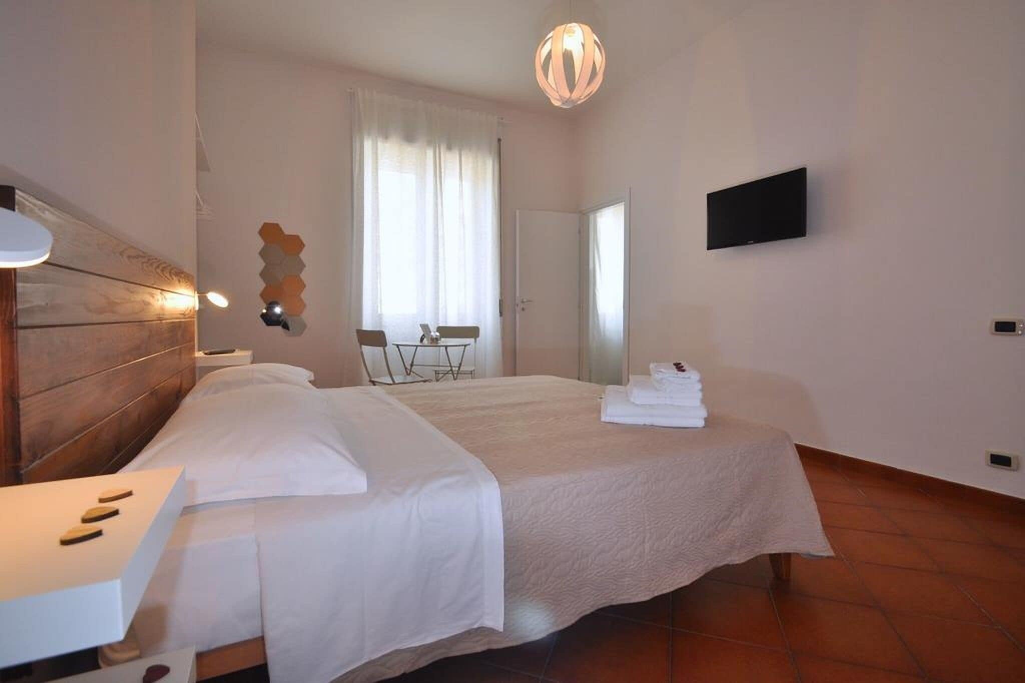 Habitación de hotel con cabecera de madera, cama con ropa de cama blanca, mesita de noche, televisor de pantalla plana en la pared y un gran ventanal con cortinas.