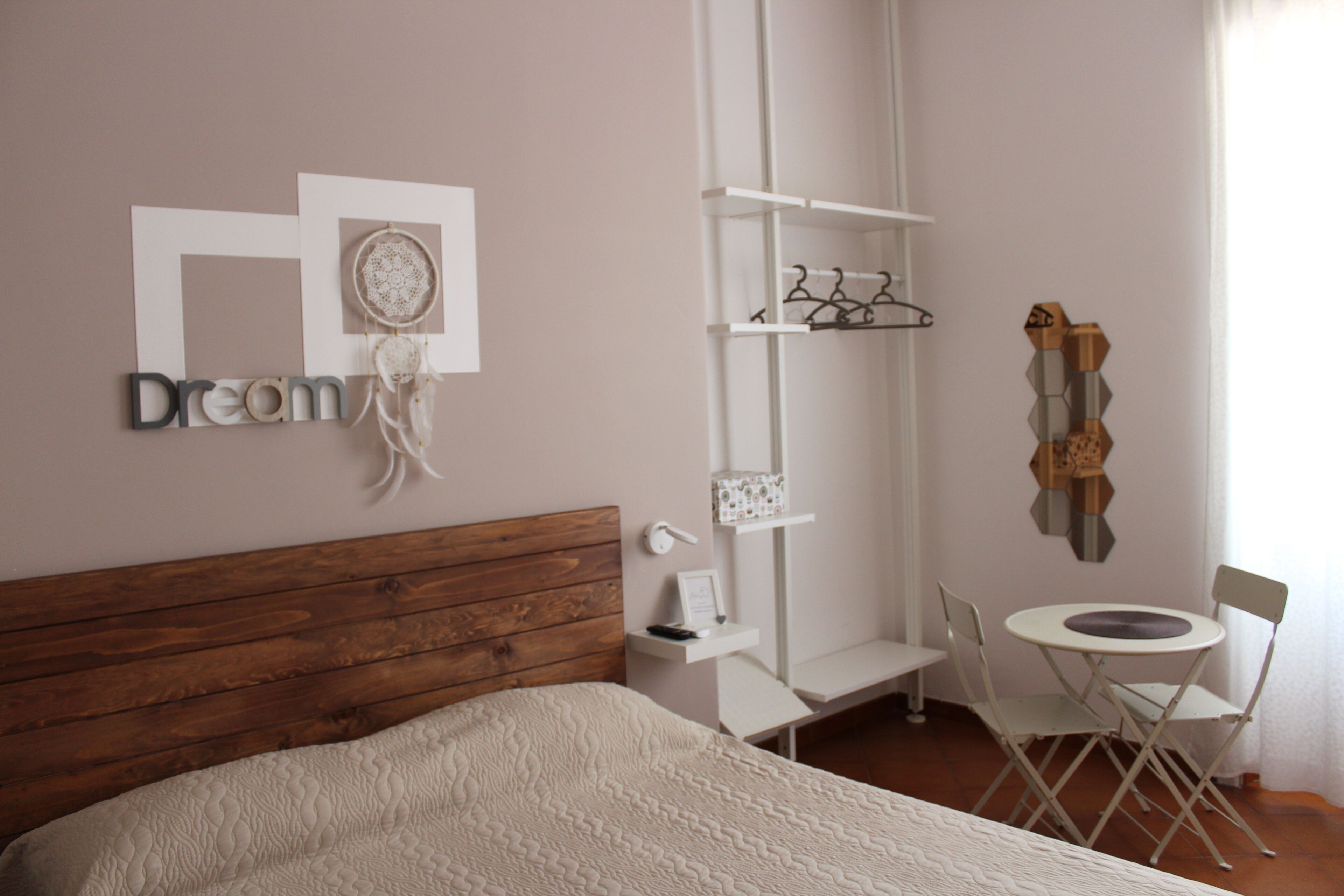 Une chambre avec une tête de lit en bois, un lit recouvert d’une couette blanche texturée, une table de chevet avec une lampe, un petit bureau blanc, une chaise, une table ronde et une étagère murale ornée d’un attrape-rêves et d’un tableau encadré.