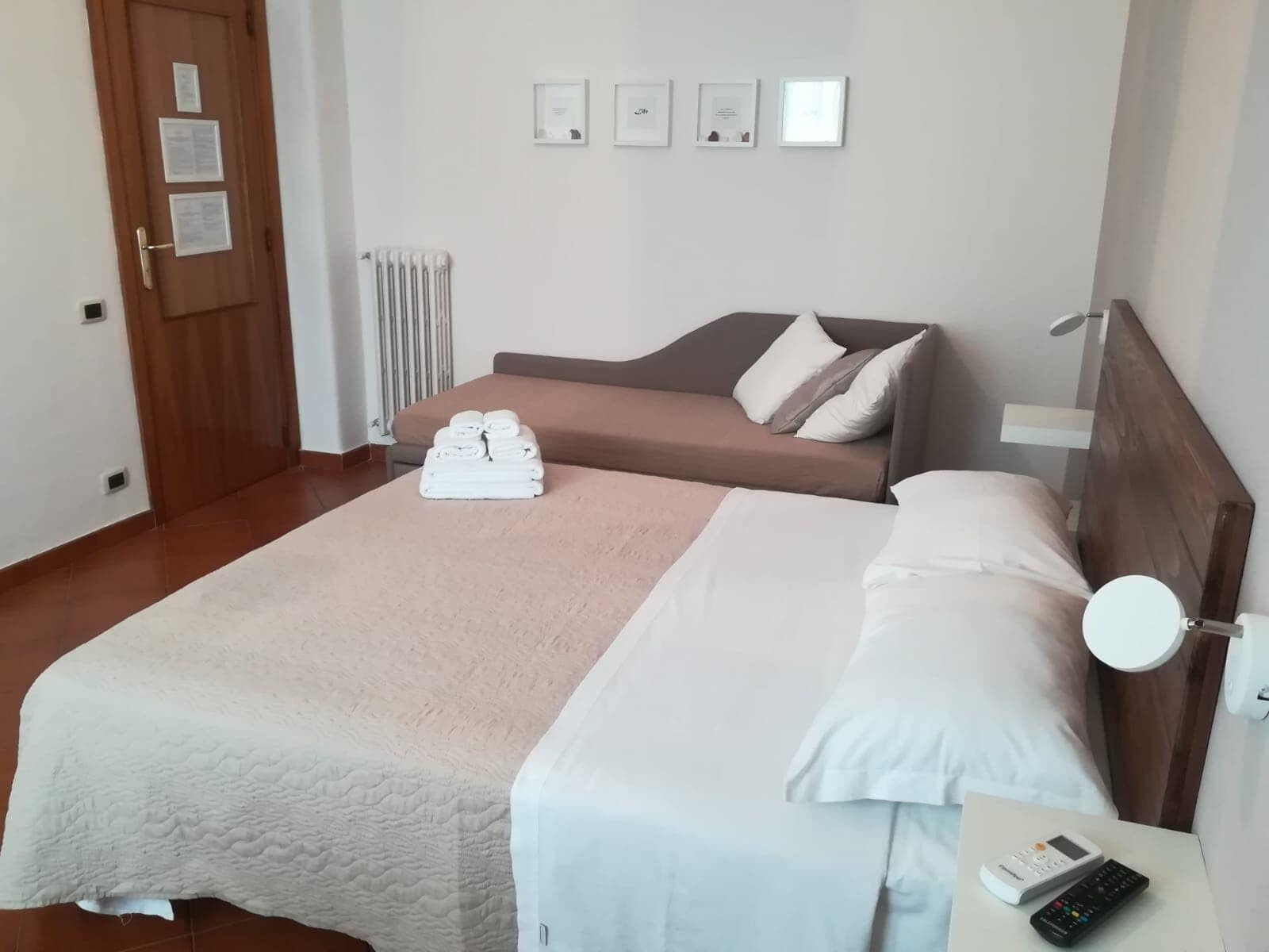 A hotel room with a bed, a bedside table with a remote control, a wall-mounted radiator, and a door with a lock.