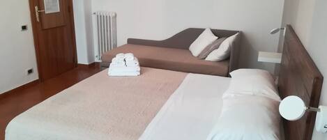 A hotel room with a bed, a bedside table with a remote control, a wall-mounted radiator, and a door with a lock.