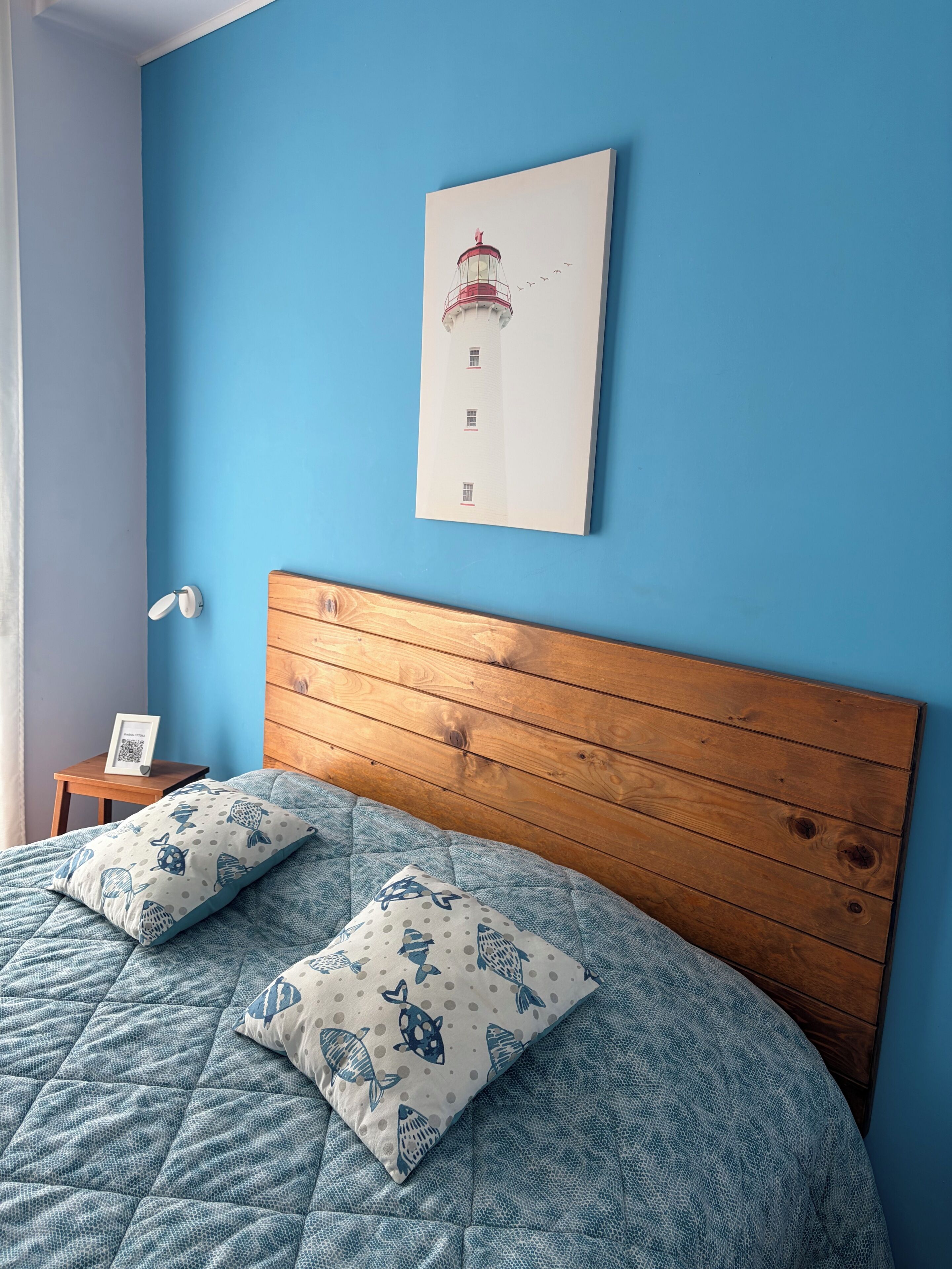 Un dormitorio con pared azul, cabecera de madera, cama con colcha azul y almohadas estampadas, un cuadro enmarcado de un faro, y una mesita de noche con una foto enmarcada y una lámpara.