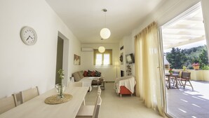 A bright, well-lit room with a dining table, chairs, a sofa, and a TV. There is a window with curtains and a view of greenery outside.