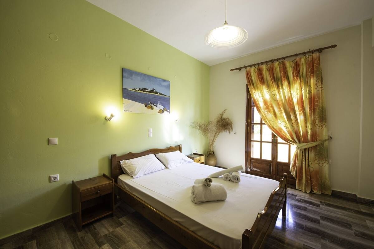 A bedroom with a bed, a nightstand, a bedside table, a wall-mounted light, a window with curtains, and a painting on the wall.