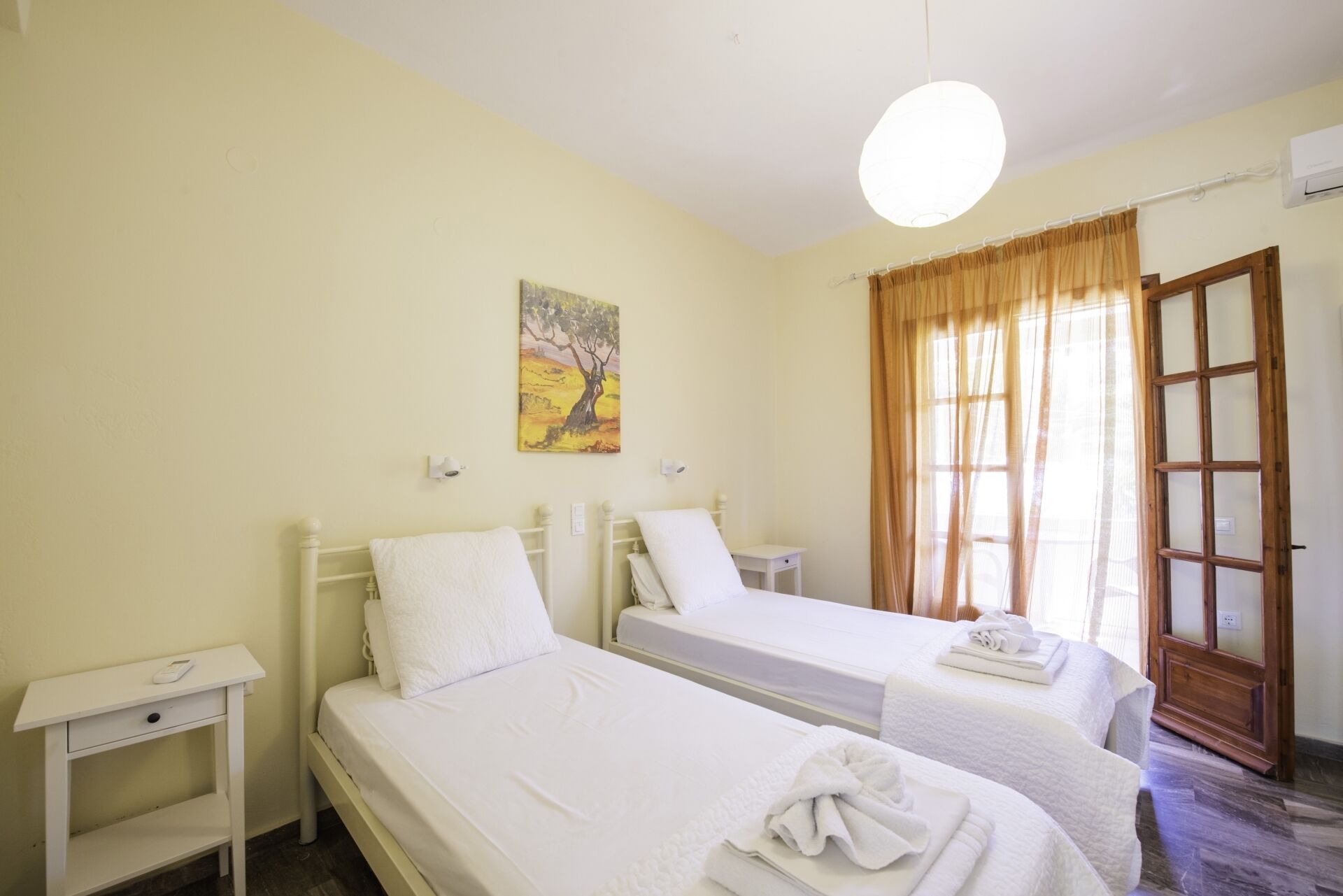 Habitación de hotel con dos camas individuales, una mesita de noche con lámpara, un cuadro en la pared y una ventana con cortinas.