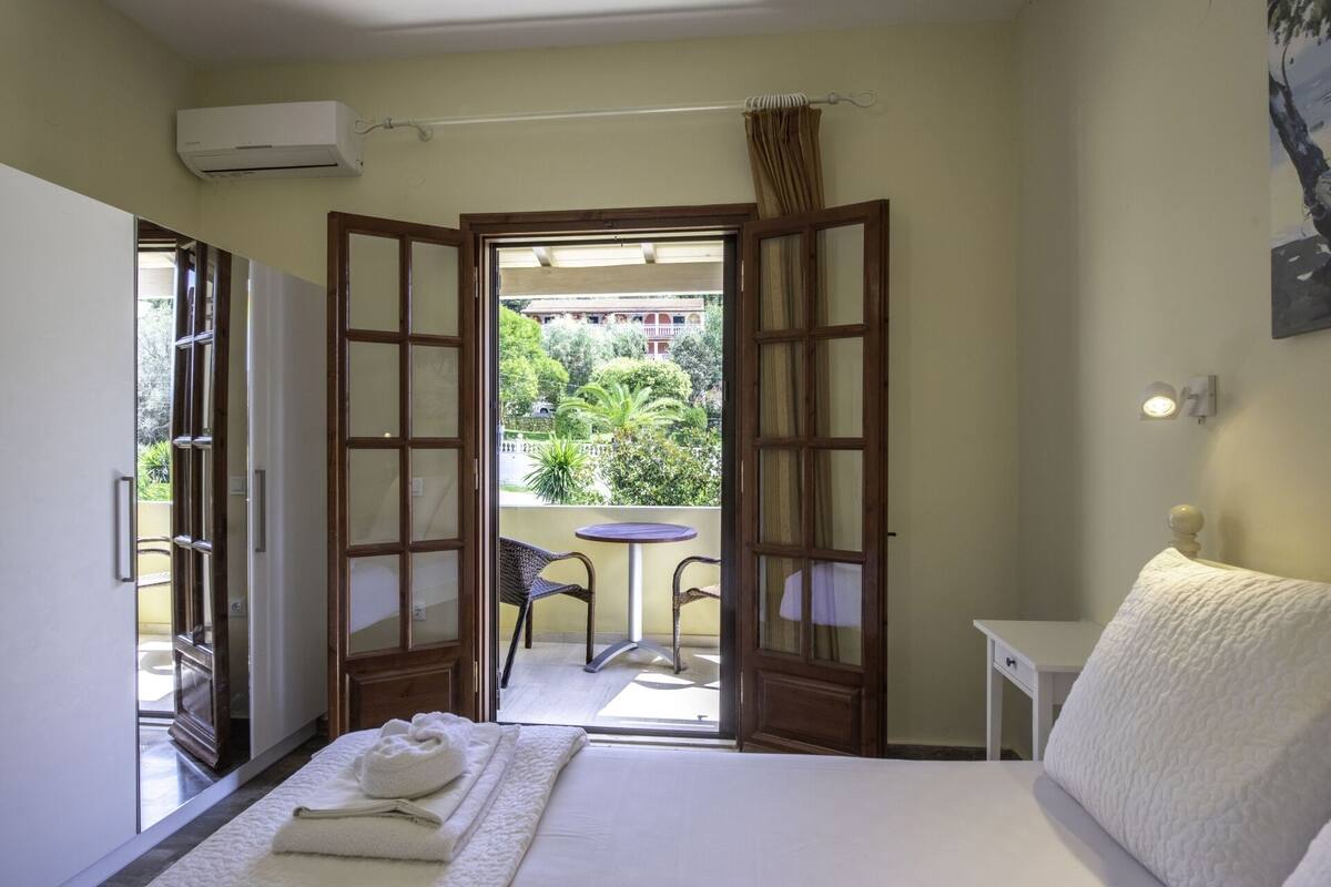 A hotel room with a bed, a small table, a chair, and an open door leading to a balcony with a view of greenery and a building.