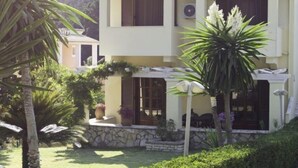 A two-story building with a stone foundation, a tiled roof, and a well-maintained garden with palm trees and hedges.