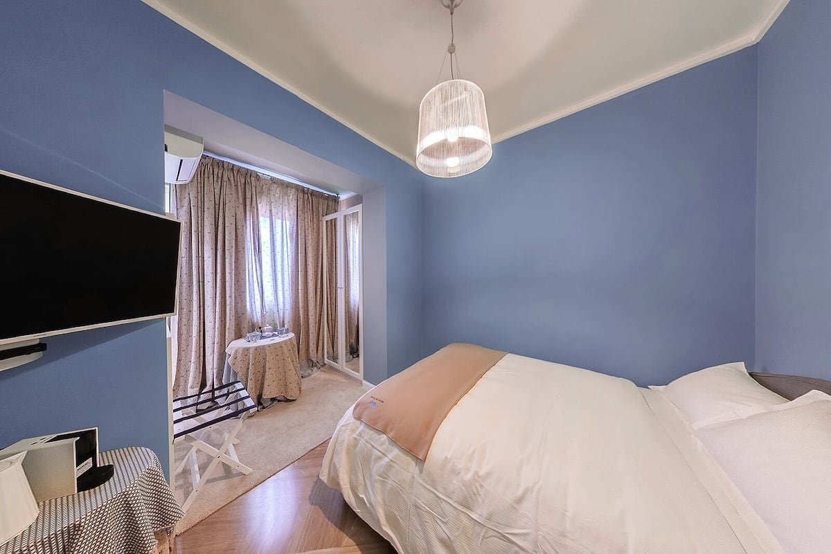 A bedroom with a bed, a TV mounted on the wall, a hanging light fixture, and a window with curtains.
