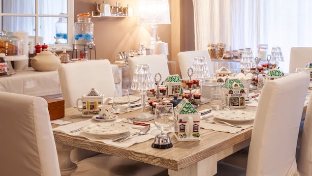 Une table à manger dressée avec de la vaisselle blanche et des décorations festives, notamment des objets en forme de maison, dans une pièce bien éclairée, dotée d’une fenêtre et de rideaux.