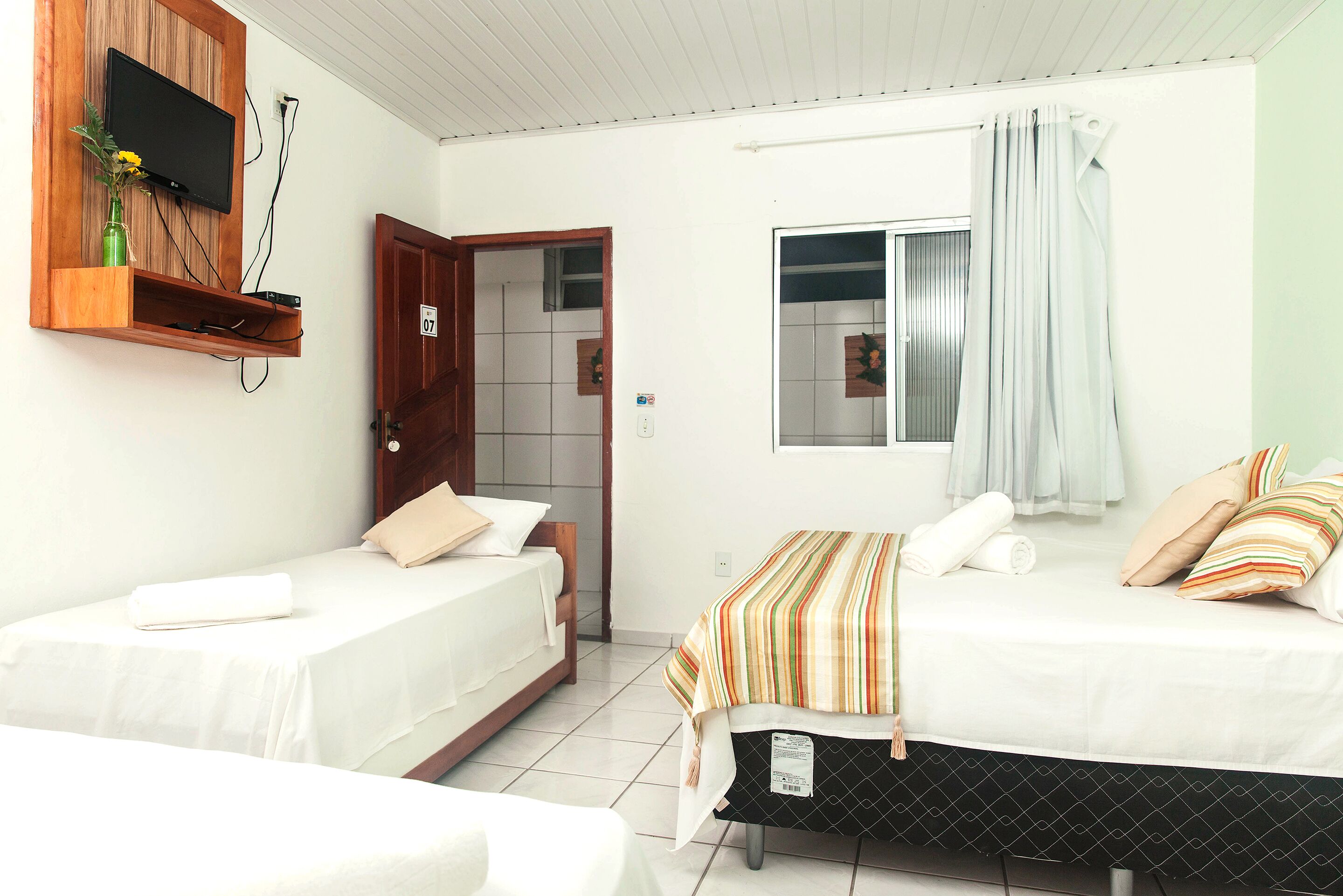 Una habitación con dos camas, un televisor montado en un estante de madera, una ventana con cortinas y una puerta.