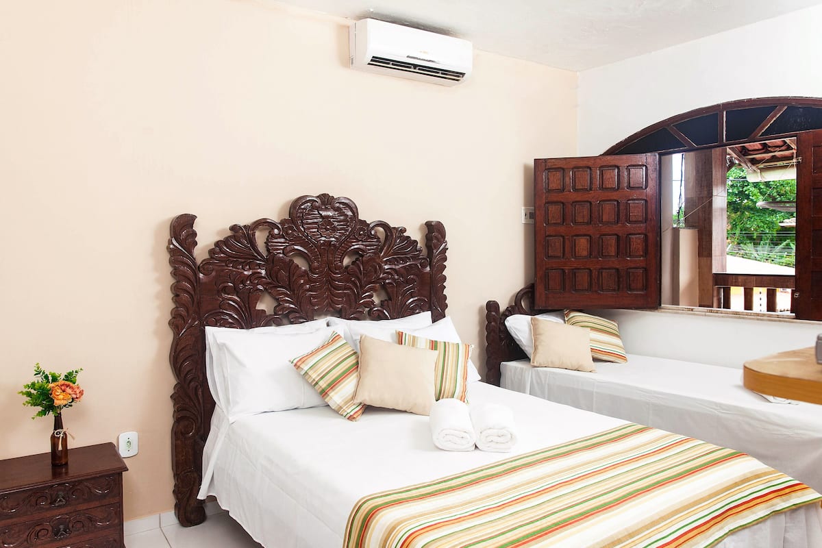 A hotel room with a wooden bed, white linens, striped pillows, and a view of greenery through an open door.