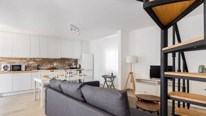 A modern kitchen with white cabinets, a dining area with a white table and chairs, a gray sofa, and a black and wood staircase. - Carvoeira Surf & Family Villa (Mafra)
