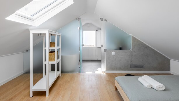 A modern, well-lit attic bedroom with a skylight, wooden flooring, a built-in white shelving unit, and a bed with a rolled-up towel. - Carvoeira Surf & Family Villa (Mafra)