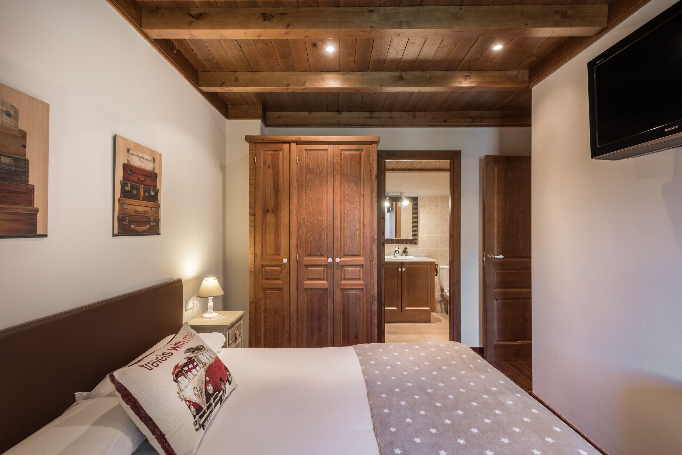 A bedroom with a wooden ceiling, a bed with a white and red pillow, a nightstand with a lamp, and a wooden wardrobe.