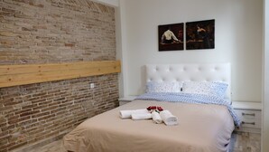 Cama bem arrumada com colcha bege e almofadas brancas, mesa de cabeceira de madeira com gaveta, parede de tijolos e duas fotos emolduradas na parede.
