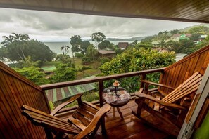 A wooden deck with two chairs and a small table, overlooking a lush landscape with houses and a body of water.