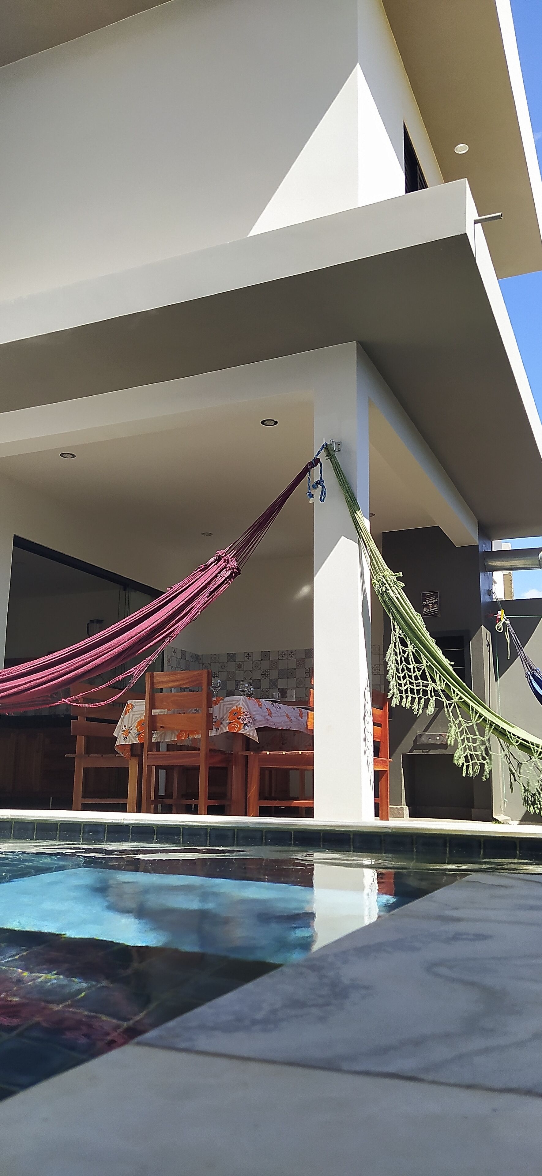 Un área de piscina con una hamaca y sillas de madera, un edificio moderno con techo plano, y un cielo despejado.