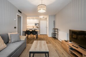 Una sala de estar moderna con un sofá gris, una mesa de centro de madera, un mueble para televisión y un comedor con una cocina al fondo.