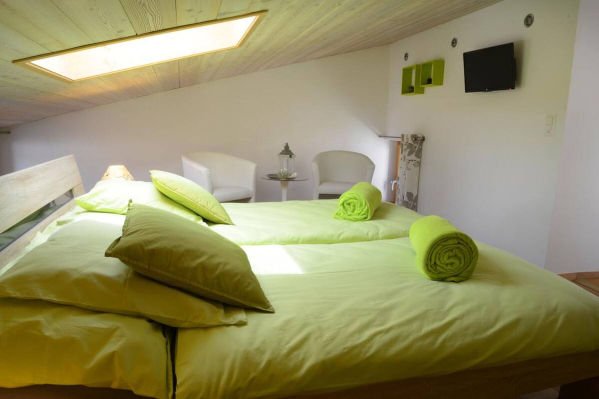 A room with a bed, two chairs, a small table with a lamp, and a wall-mounted TV.
