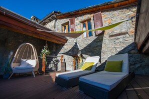 A stone building with a wooden deck, a hanging chair, and two lounge beds with pillows. - Chez Constance (Saillon)