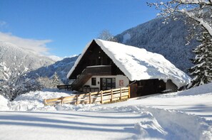 Una cabaña de madera con techo cubierto de nieve, rodeada de árboles y montañas nevadas.