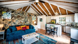 Ein geräumiger Loft mit hoher Decke, freiliegenden Holzbalken, einer Steinwand, einem blauen Sofa, einem Essbereich und einer Küche.