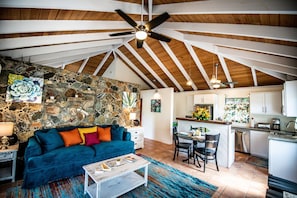 A spacious loft with a high ceiling, exposed wooden beams, a stone wall, a blue sofa, a dining area, and a kitchen. - Salty Kisses & Goodnight Wishes-Come enjoy St. John, USVI, in peaceful Coral Bay (St. John)