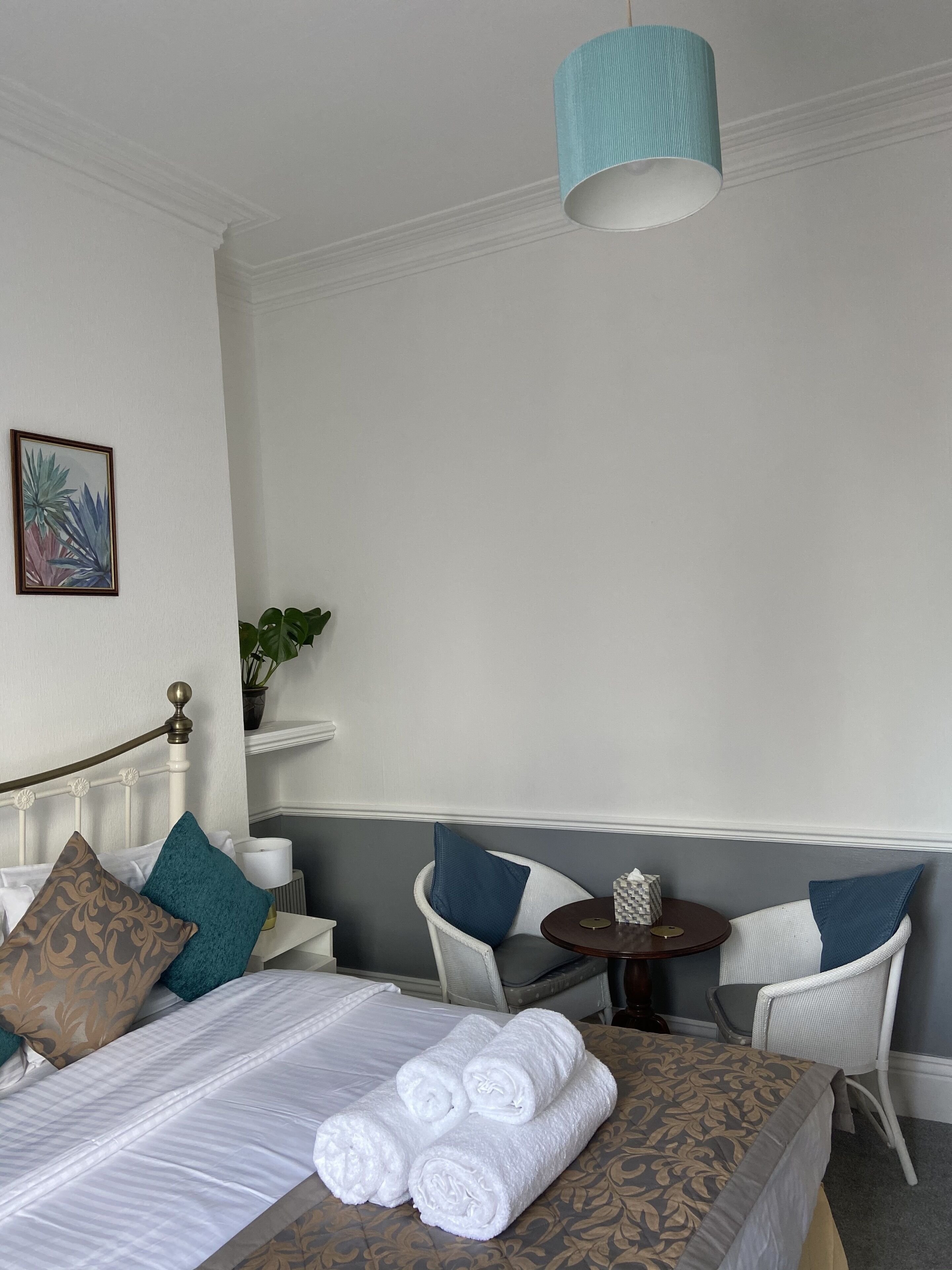 Une chambre d’hôtel avec un lit soigneusement fait, deux chaises blanches, une petite table ronde avec un boîte de mouchoirs et un luminaire suspendu.