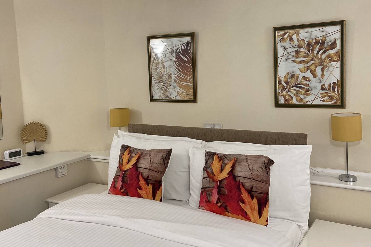 A hotel room with a bed, two pillows with autumn leaf designs, a bedside table with a lamp, and two framed artworks on the wall.