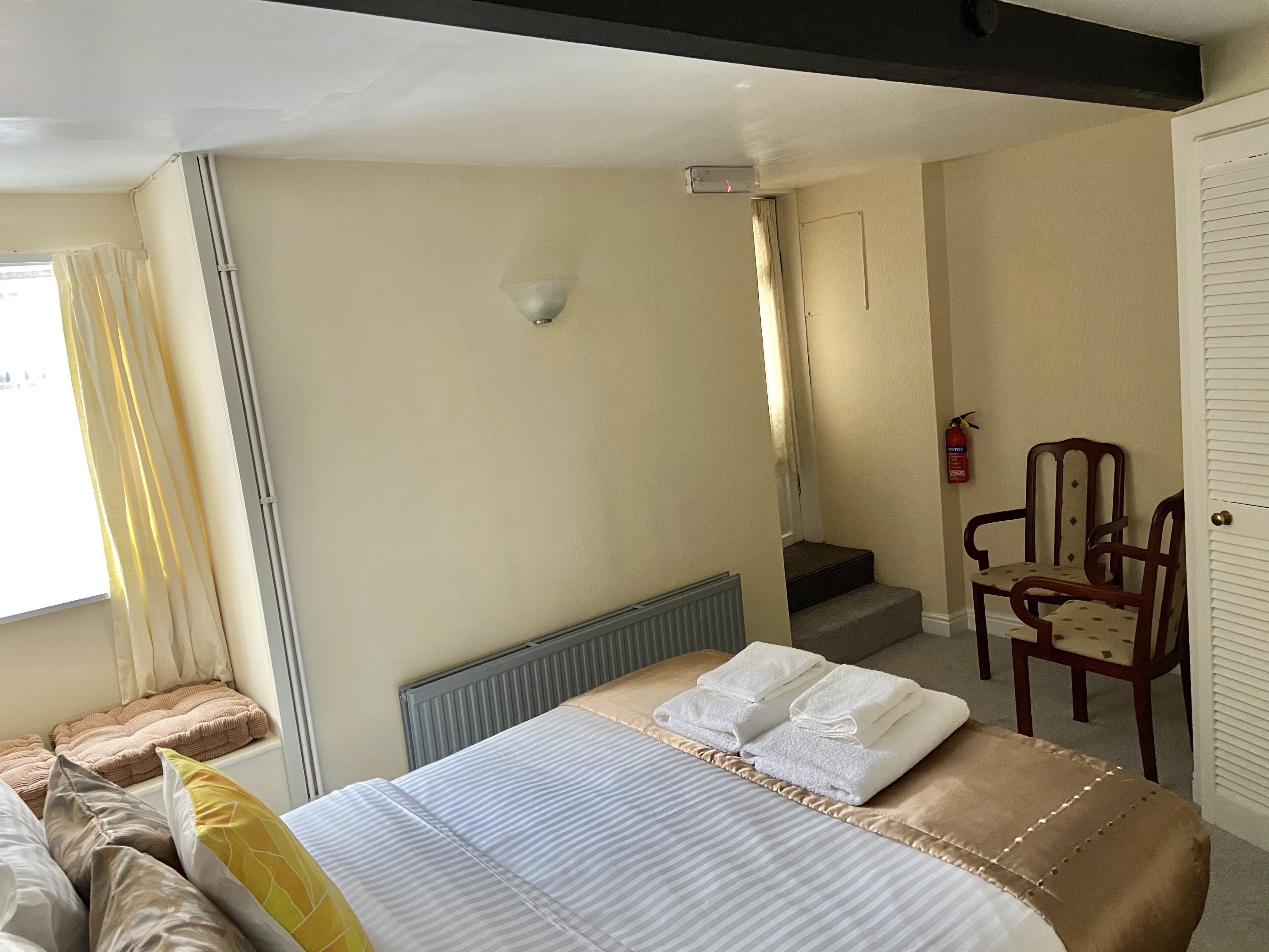 Habitación de hotel con cama, una silla, radiador, ventana con cortinas, puerta y escalera.