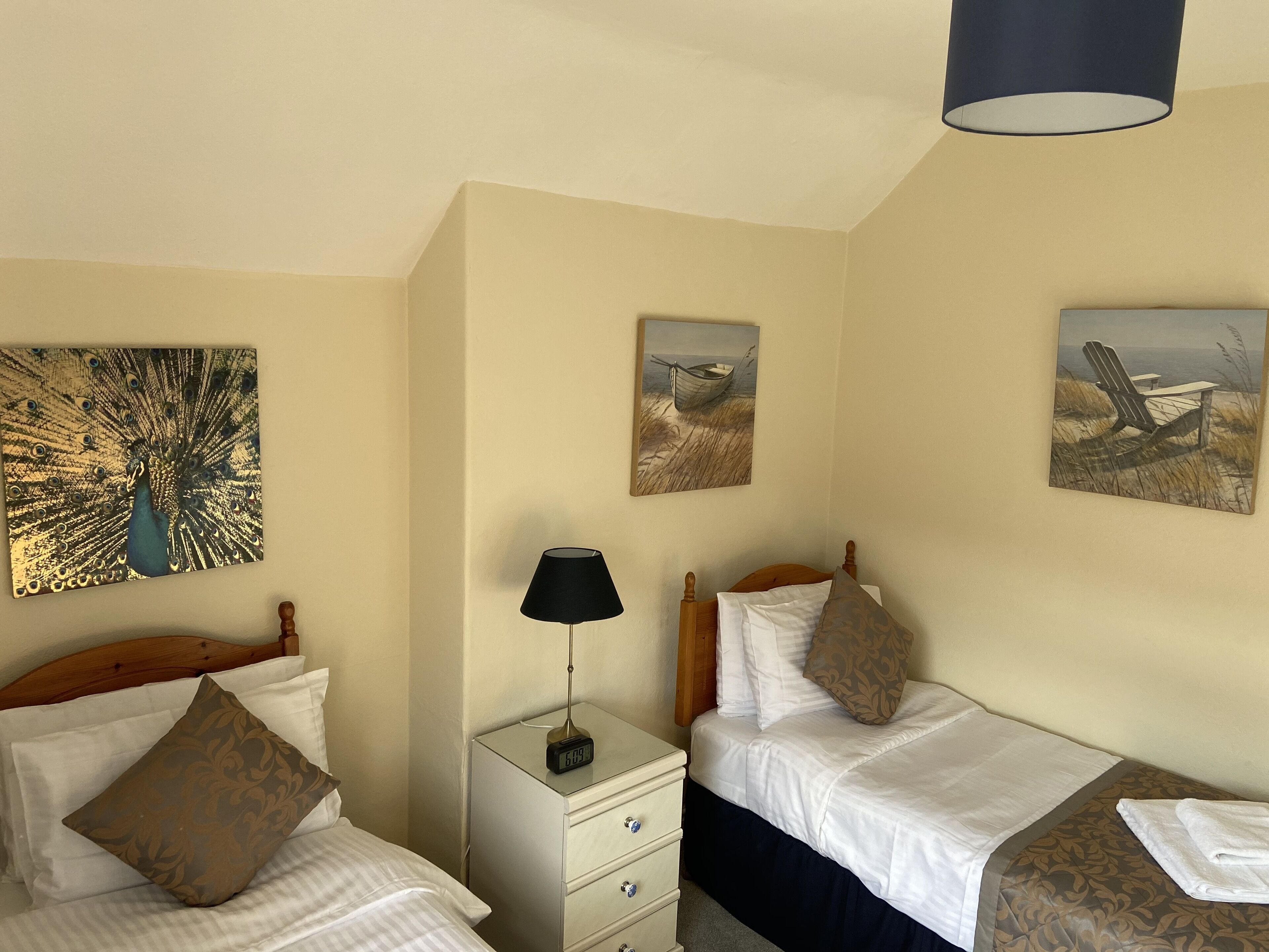 A hotel room with two single beds, a nightstand with a lamp, and two wall-mounted paintings.