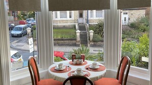 A dining area with a round table set for four, two chairs, and a view of the street and garden through large windows.