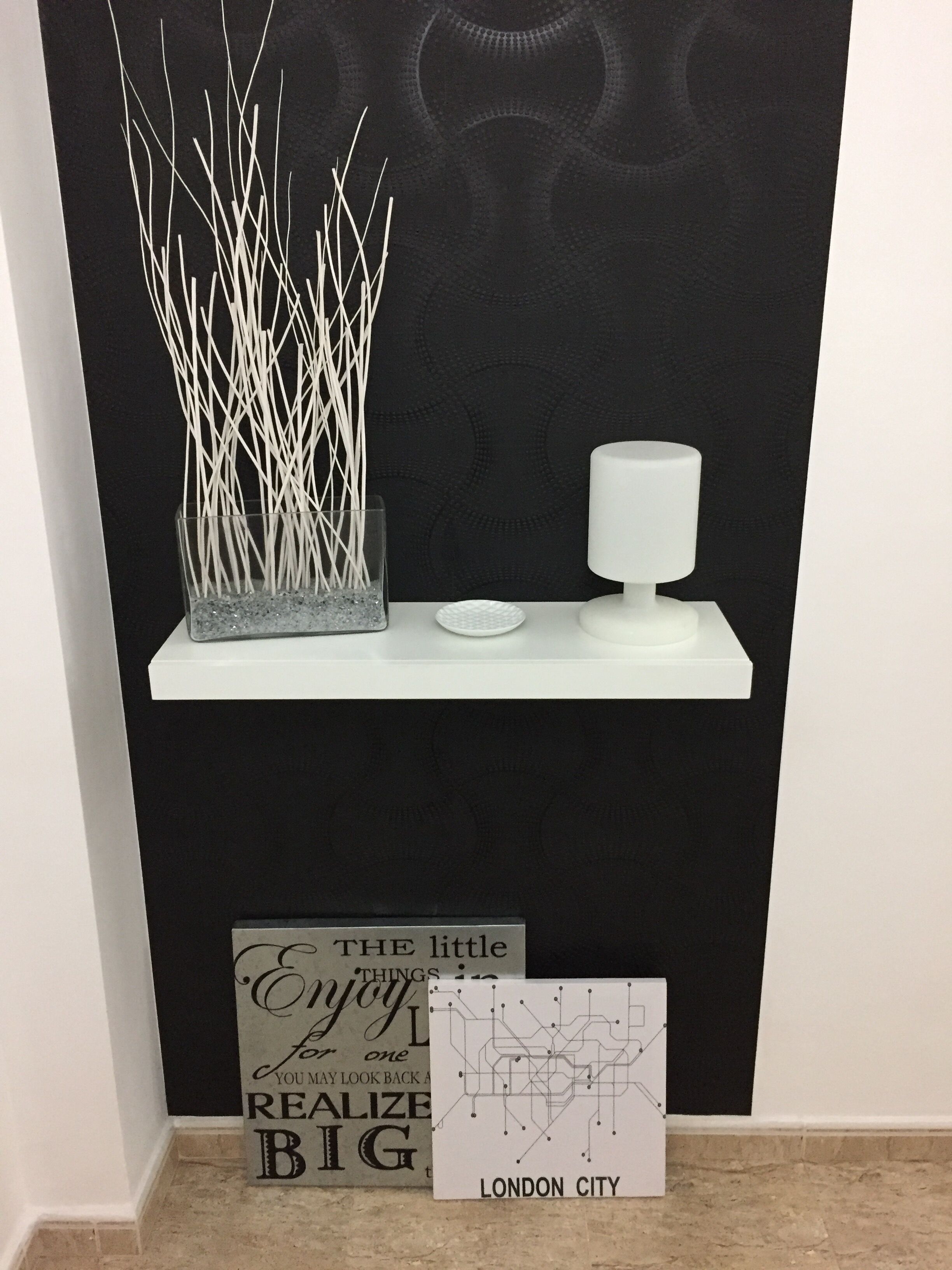 Eine schwarze Wand mit einem weißen Regal, das eine weiße Vase und ein kleines, weißes Objekt beherbergt. Darunter befinden sich zwei Schilder, eines mit dem Text 'THE little ENJOY for one YOU MAY LOOK BACK BIG' und ein weiteres mit 'LONDON CITY' und einer Karte.