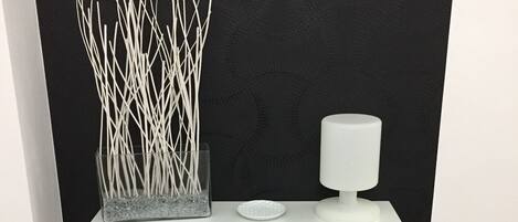 Eine schwarze Wand mit einem weißen Regal, das eine weiße Vase und ein kleines, weißes Objekt beherbergt. Darunter befinden sich zwei Schilder, eines mit dem Text 'THE little ENJOY for one YOU MAY LOOK BACK BIG' und ein weiteres mit 'LONDON CITY' und einer Karte.