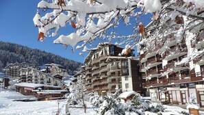 Une station de montagne enneigée, avec des bâtiments à plusieurs étages, un télésiège et un ciel bleu dégagé.