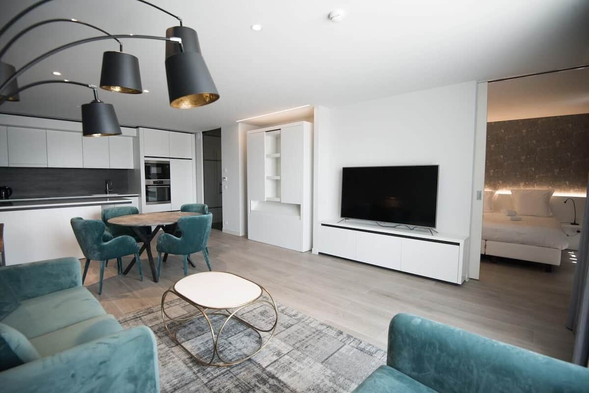 A modern living room with a flat-screen TV, teal sofas, a dining area with a round table, and a kitchen with white cabinets.