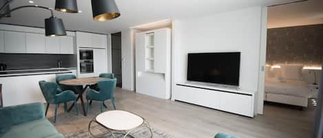 Una sala moderna con televisor de pantalla plana, sofás color turquesa, un comedor con mesa redonda y una cocina con armarios blancos.