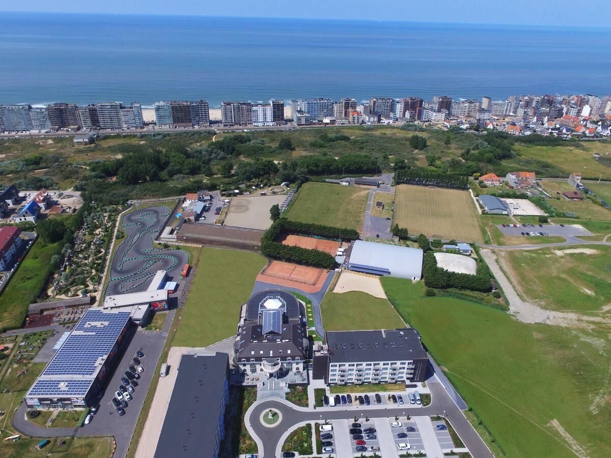 An aerial view of a coastal area with a large building complex, including a sports field, parking areas, and a solar panel installation.