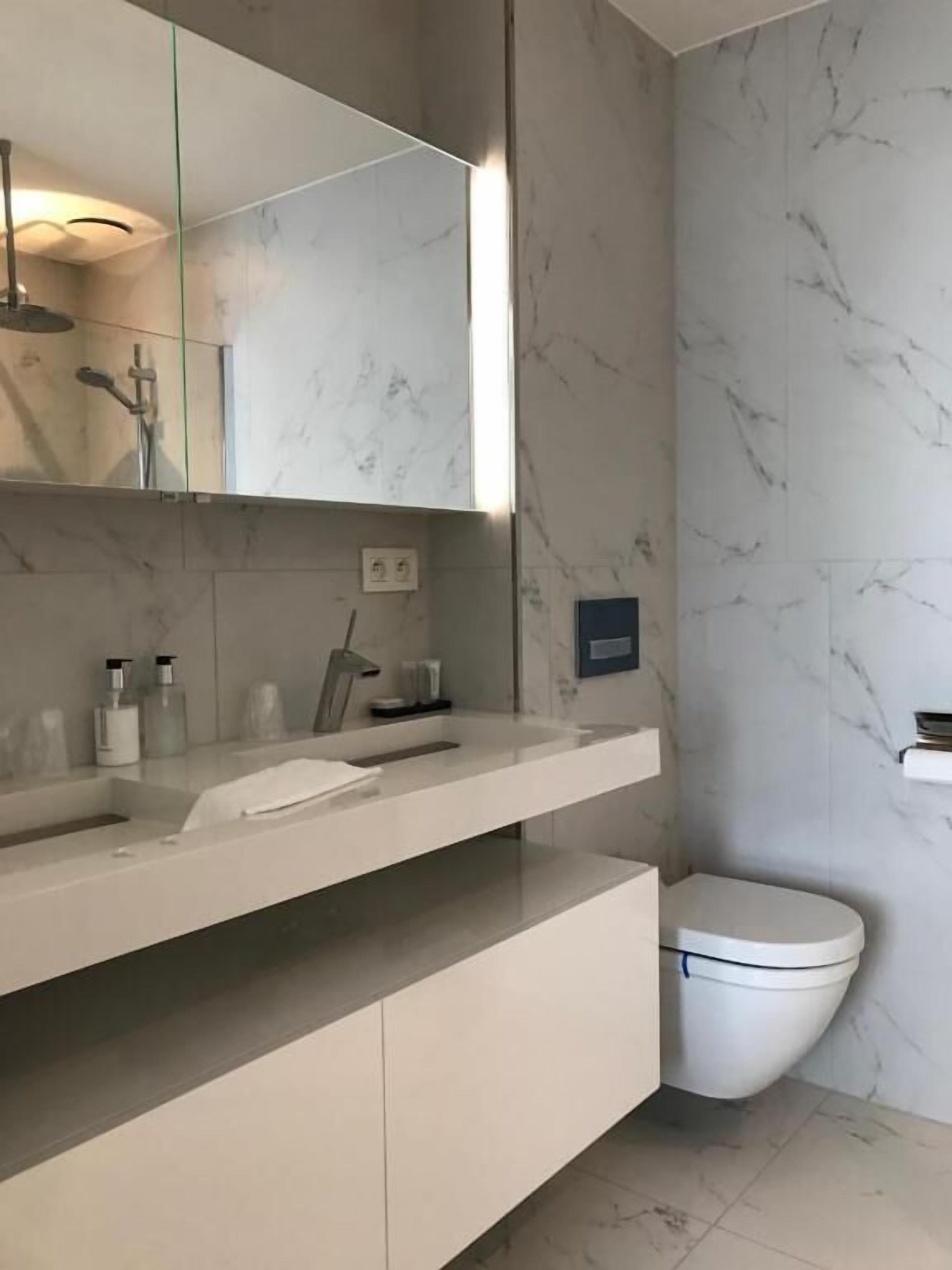 A modern bathroom with a marble backsplash, a large mirror, a white sink, and a toilet.