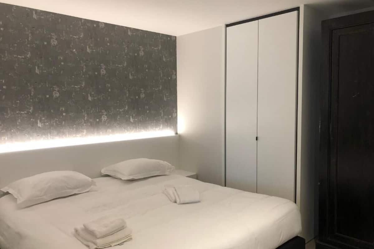 A modern bedroom with a large bed, white linens, a headboard with integrated lighting, and a dark wardrobe.