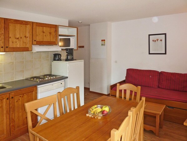 A compact kitchen with wooden cabinets, a microwave, a coffee maker, and a dining table with chairs. There is a red sofa in the living area. - Cozy 2-Room Apartment for 6 with Pool, Jacuzzi, Sauna, and Mountain Views (Les Orres)