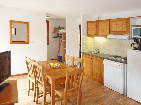 A compact kitchen with wooden cabinets, a built-in dishwasher, and a dining area with a wooden table and chairs. - Cozy 2-Room Apartment for 6 with Pool, Jacuzzi, Sauna, and Mountain Views (Les Orres)