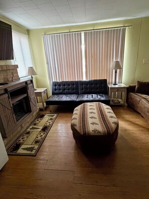 A living room with a sofa, ottoman, fireplace, and blinds. - Close to Tippy Dam, Lake Elinor, Dickson Park. Clean & cozy! (Brethren)