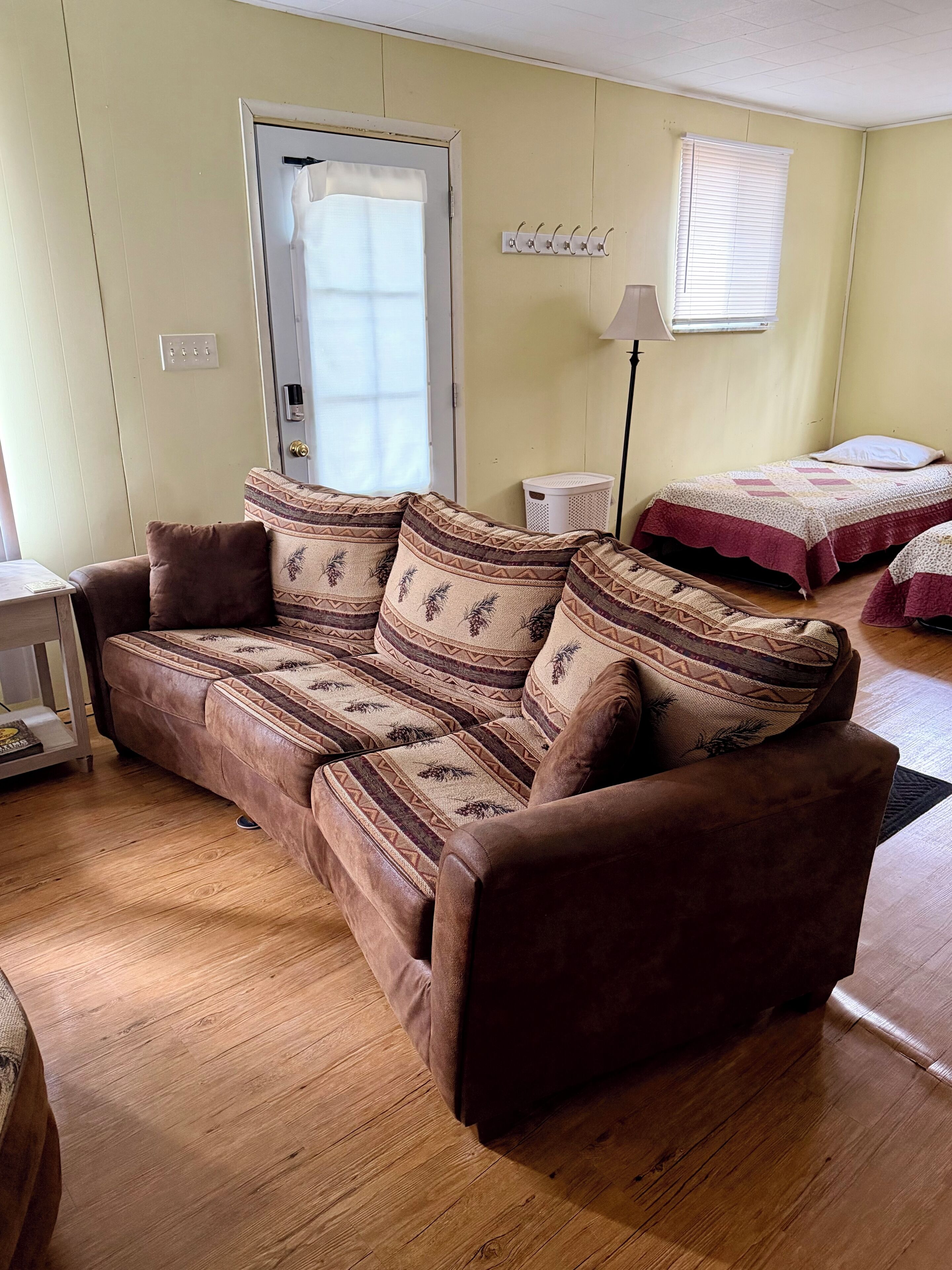 Une pièce avec un canapé marron, un lit recouvert d’une couvre-lit à motifs, une lampe et une fenêtre avec des stores.